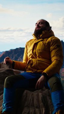 Dikey Video Erkek dağ yürüyüşçüsü muzaffer bir tırmanışı kutluyor ve kolları heyecan içinde kayaların üzerinde otururken dinleniyor. Manzaralı bir arka planda panoramik manzara ve temiz hava