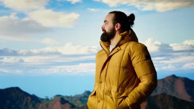 Sıcak kıyafetler giymiş yürüyüşçü zirveye tırmandıktan sonra bir dağın zirvesinde duruyor, engebeli tepelerin ve engin gökyüzünün nefes kesici manzarasına bakıyor. Manzaralı macera. Kamera B.