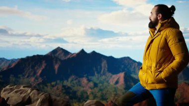 Sıcak kıyafetler giymiş yürüyüşçü zirveye tırmandıktan sonra bir dağın zirvesinde duruyor, engebeli tepelerin ve engin gökyüzünün nefes kesici manzarasına bakıyor. Manzaralı macera. Kamera A.