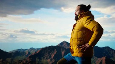 Doğa aşığı nefes kesen bir dağ sırasının kenarında duruyor, yükselen zirvelerin ve uçsuz bucaksız gökyüzünün manzaralı arka planına bakıyor. Macera ve açık hava keşiflerini seven genç bir adam. Kamera A.