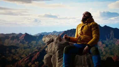 Erkek dağcı muzaffer tırmanışını kutluyor ve kolları heyecan içinde kayalarda otururken zirvede dinleniyor. Manzaralı bir arka planda panoramik manzara ve temiz hava. Kamera A.