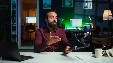 İçerik yaratıcısı cep telefonuyla konuşuyor stüdyoda selfie çubuğuna bağlı, mikrofon kullanarak izleyicilerle sohbet ediyor. Telefon kamerası ve profesyonel mikrofon kullanarak vlog, kamera B 'yi kaydeden adam.