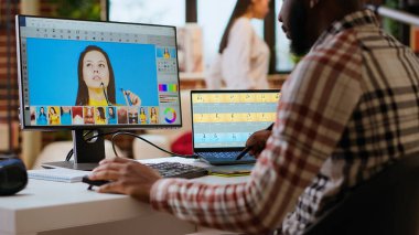 Afrikalı Amerikalı erkek tasarımcı çift ekran görüntülerini düzenliyor, yaratıcı bir ofiste bilgisayar kurulumu yapıyor. Dijital medya profesyoneli için fotoğraf rötuşlamaya odaklı ideal çalışma alanı. Kamera B.