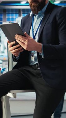 Dikey video Photovoltaics fabrikası çalışanı, güneş panellerini montaj hatlarına yerleştiren robotların performansını gözden geçirmek için tablet kullanıyor. Masada oturan uzman, güneş santralini incelemek için aygıt kullanıyor.