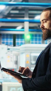 Fotovoltaik fabrikasındaki dikey video adam dizüstü bilgisayardaki izleme sistemi performansını gösteriyor. Güneş panelleri enerji üretimini ve verimliliğini izlemek için dizüstü bilgisayar kullanan fabrika çalışanı üretiyor.