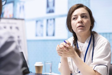 Hekim, bir kabinede tıbbi sonuçları tartışmak için bir hastayla buluşur. Röntgen taramalarını yorumlamak için gelişmiş teşhis ekipmanları kullanır ve profesyonel bir sağlık hizmeti yaklaşımı gösterir..