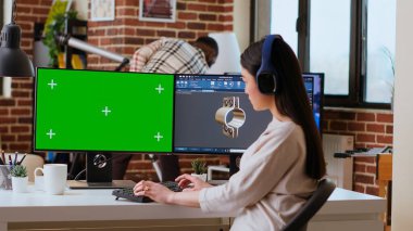 Yazılım geliştiricisi, yeşil ekran ve çift monitörlü modern bir ofiste çalışıyor. Ev işleri endüstriyel yenilikleri ve imalat alanında uzmanlığı vurguluyor. Kamera B.