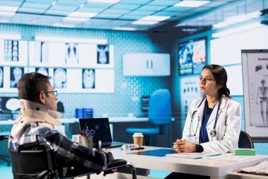 Hintli doktor, tekerlekli sandalyedeki hastaya röntgen sonuçlarını açıklıyor. Fizyoterapi öneriyor ve profesyonel tavsiyelerde bulunuyor. Kadın doktor boyun tasmalı adama yardım ediyor..