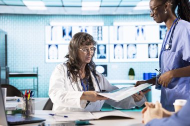 Hekim, Afrikalı Amerikalı hemşirelerden kabinedeki hasta dosyalarını istedi. Karşılaştırma için eski test sonuçlarını içeren sağlık raporlarını okudu. Tıbbi ekip yaşlı bir adamla taramaları inceliyor..