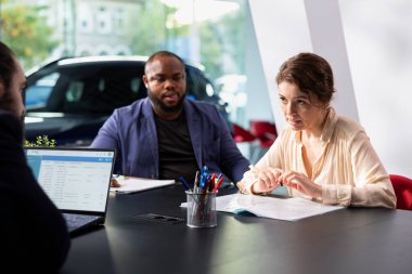 Satış temsilcisi, müşteriler için araba test sürüşü planlamak için dizüstü bilgisayardaki randevu listesini inceliyor. Showroom temsilcisi müşterilerle tartışıyor, test sürüşünü düzenliyor