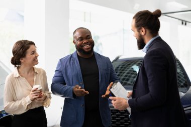 Gülümseyen satıcı mutlu müşterilerle konuşuyor, galeri araçlarını gösteriyor ve onların özelliklerini anlatıyor. Neşeli müşterilere araba özelliklerini sunan neşeli bir galeri temsilcisi.