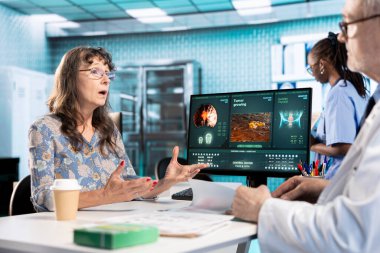 Kıdemli bayan hasta doktoruna yeni bir tedavi planı soruyor, sağlık kayıtlarını ve tarama sonuçlarını değerlendiriyor. Tıbbi uzman, hastaya iyileşme yolunda rehberlik ediyor..