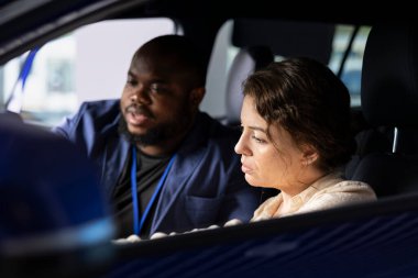 Araba galerisi satıcısı test sürüşü sırasında müşteriyle konuşuyor, mevcut modeller hakkında bilgi sağlıyor. Afrikalı Amerikalı ajan müşteriyle araç fiyatları pazarlığı yapıyor