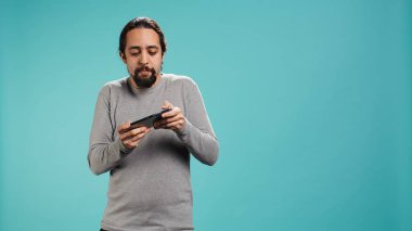 Odaklanmış adam cep telefonuyla yoğun video oyunu oynuyor, dokunmatik ekrandaki saldırı tuşlarını spamlıyor. Oyuncu oyun yarışmasına katılıyor, stüdyo arka planında izole edilmiş, kamera B