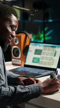 Vertical Video Black serbest çalışanı online verileri araştırıyor ve makaleler için notlar yazıyor, kişisel ofisinde oturuyor. Evden çalışan genç bir adam, kablosuz bir kurulum kullanarak görevleri dengeliyor. Kamera.