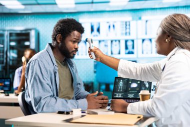 Siyahi pratisyen, tesiste rutin kontroller sırasında ateşi ölçerek grip belirtilerini dijital termometreyle kontrol ediyor. Sağlık hekimi genç bir hastaya teşhis koyuyor..