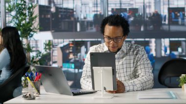 Siyahi bir çalışan ofis masasında telekomünikasyon toplantısına katılıyor, proje yönetimi ve iş stratejisi tartışmaları arasında birden fazla iş yapıyor. Bölümler arası verimlilik ve işbirliği. Kamera A.