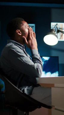 Ev işyerinde esneyen migreni olan dikey video genç zenci çalışan, yeni yönetim görevlerini çözmek için online raporları inceliyor. Afrikalı Amerikalı adam projeden dolayı yorgun ve stresli.