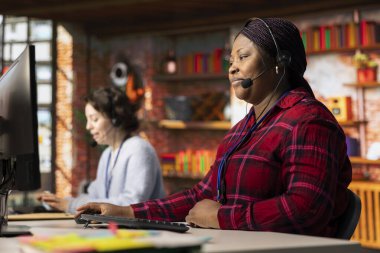 Çağrı merkezi çalışanı, yazılımla ilgili teknik sorulara cevap vermek için ses kulaklığı kullanıyor. Afrika kökenli Amerikalı bir kadın müşteri hizmetlerinde sorun çözmek için bilgisayar kullanıyor.