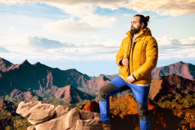 Sıcak kıyafetler giymiş yürüyüşçü zirveye tırmandıktan sonra bir dağın zirvesinde duruyor, engebeli tepelerin ve engin gökyüzünün nefes kesici manzarasına bakıyor. Manzaralı macera.