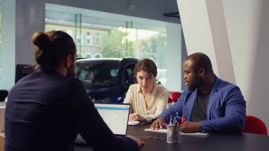 Galerideki müşteriler araba satışıyla ilgili gerekli ödeme belgelerini tamamlıyorlar, düzgün işlemler için finans ve sigorta departmanlarıyla işbirliği yapıyorlar, kamera B