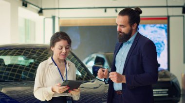 Satıcı kadın, araba seçimini erkeklere göstermek için tablet kullanıyor. Satın alması için onu ikna etmeye çalışıyor. Showroom temsilcisi anlaşma müşterisi sunuyor, cihazda ayrıntıları gösteriyor, satış taktikleri kullanıyor, kamera A