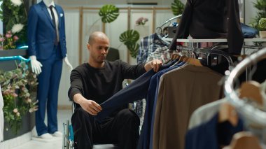 Tekerlekli sandalyedeki bir ortadoğulu müşteri perakende mağazasında yeni giysiler, tüketim ve stil seçimi inceliyor. Kronik maluliyet sorunu olan genç bir adam moda ürünler için alışveriş yapıyor. Kamera A.