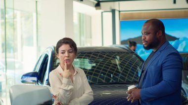 Araba galerisinde yürüyen müşteriler mevcut modellere göz atıyorlar ve ideal yatırım seçeneğini seçmeye çalışıyorlar. Galeride araç almak isteyen karı koca, kamera A.