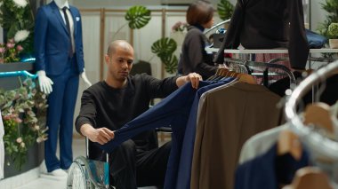 Tekerlekli sandalyedeki erkek kullanıcı, engelli dostu bir butikte yeni moda kıyafetler arıyor. Kıyafet alışverişinin keyfini çıkarıyor. Orta Doğulu bir adam kişisel tarzını yükseltiyor. Kamera A.