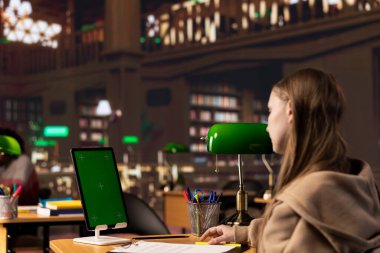 Üniversite öğrencisi güvenilir akademik kaynaklardan notlar toplarken tabletteki bir fotokopi ekranına bakıyor, bir ödev için kampüs kütüphanesinin kitap koleksiyonuna danışıyor. Öğrenci hayatı.