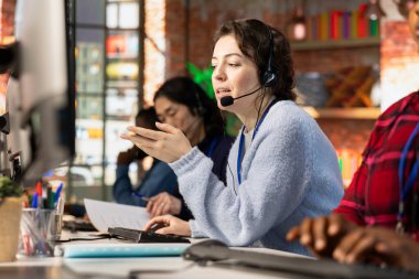 Çağrı merkezi operatörü üzgün müşterilerle ilgilenen iş arkadaşlarının yanında ofis masasındaki kulaklıkla konuşuyor. Müşteri desteği işi yapan ekip çalışanları, müşteri şikâyetleriyle ilgileniyorlar