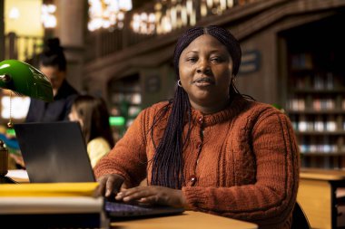 Akademik kaynaklar ve kütüphane veritabanında öğrenme materyalleriyle çalışan Afrikalı Amerikan kütüphanecisinin portresi. Gülümseyen kadın eğitim mesleğine odaklanmış..