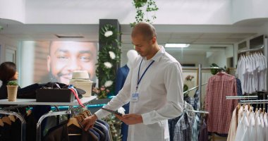 Orta Doğulu perakende işçisi raflardaki yeni ürün koleksiyonunu düzenliyor, şık bir alışveriş merkezinde moda ürünleri düzenliyor. Tezgahtar detaylara dikkat ediyor. Kamera B.