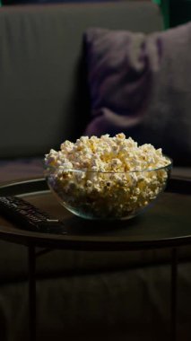 Loş ışıklandırmalı, patlamış mısırlı ve uzaktan kumandalı dikey video odası film gecesi için rahat bir ortam yaratıyor, kapatın. Oturma odasındaki masada patlamış mısır kasesi.