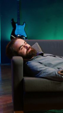 Dikey videodaki gülümseyen adam evde kanepede uzanmış patlamış mısır yerken televizyon kanalında yayınlanan filmlere göz gezdirip işten izin almanın keyfini çıkarıyordu. Apartmanda uzanmış yemek yiyen bir adam.