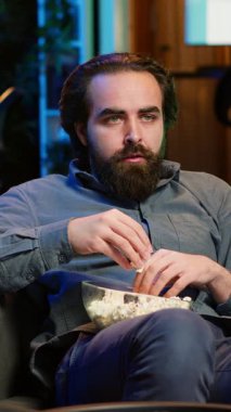 Evde dikey videocu TV 'deki gerilim filmine güvensizlik içinde bakıyor, ağzına patlamış mısır tıkıştırıyor. VOD kanalında gerilimli film izleyen şaşkın insan, şaşırmış durumda.
