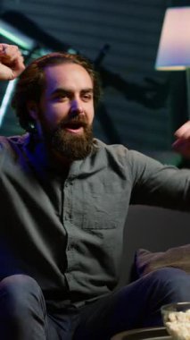 Dikey video ekstatik spor hayranı evde canlı yayında maç seyrediyor ve favori takım golünü kutlamak için bağırıyor. Öforik adam bağırıyor, el kol hareketi yapıyor, gol ve kameraya bayılıyor.