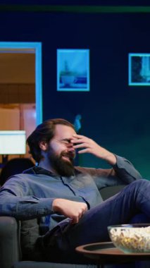 Dikey video şen şakrak bir adam kanepede oturmuş komik komedi filmleri seyrediyor, patlamış mısır yiyor. Apartmanda atıştırmalık yiyen ve film sahnesinde komik bir şakayla eğlenen neşeli bir insan.