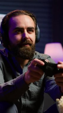 Dikey video tedirgin adam video oyunları oynuyor, kontrol cihazını kullanıyor, neon ışıklı odada eğleniyor. Coşkulu oyun kurucu oyun platformu kullanarak çok oyunculu bir oyunda yarışıyor, arkadaşlarıyla konuşuyor