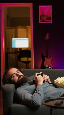 Dikey video oyuncular kanepeye uzanmış oyun alanı kullanarak çok oyunculu oyun oynuyorlar, atıştırmalık şeyler yiyorlar. Ev sinemasında oyun konsolunda kucağında patlamış mısırla video oyunu oynayan kişi, kamera B