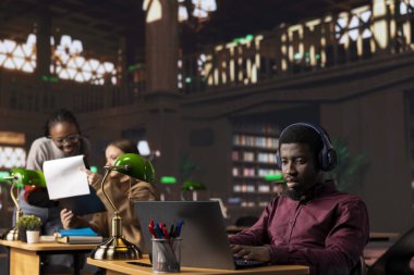 Afrikalı Amerikalı erkek öğrenci, kampüs kütüphanesinde kulaklıkla bir web konferansına katılıyor, notlar alıyor ve geleneksel olmayan bir canlı etkinlikte politika hakkında bilgi ediniyor. Adam akademik uzmanları dinliyor..