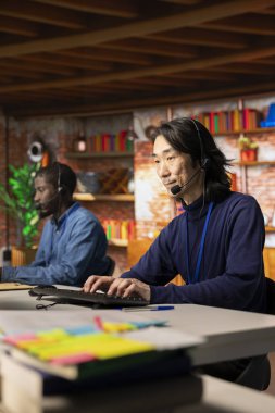 Tecrübeli çağrı merkezi kıdemli ajanı mikrofonla konuşuyor, müşterilerin hesaplara ve cihazlara tekrar erişmelerine yardım ediyor. Müşteri merkezindeki Asyalı adam parolayı sıfırlamak için yardıma ihtiyacı olan müşterilere yardım ediyor