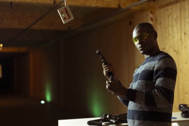 Atış poligonunun portresi. Müşteri silahını dolduruyor. Hedefleri vurmaya hazırlanıyor, eğleniyor. Kapalı alanda ateş eden, silah dolduran, koruyucu gözlük takan bir adam.