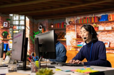 Ofisteki kıdemli çağrı merkezi temsilcisi müşterilere sorulara cevap vererek, sorunları çözerek yardımcı oluyor. Müşteri destek çalışma alanında deneyimli bir Asyalı müşterilerle konuşmak için kulaklık mikrofonunu kullanıyor.