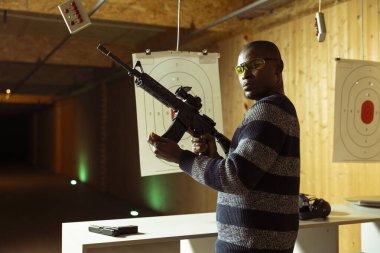 Atış poligonundaki müşterilerin tüfeklerini doldurup boş zamanlarında hedef vurmaya hazırlandıkları portre. Kapalı alanda ateş eden adam yarı otomatik silah yüklüyor, koruyucu gözlük takıyor.