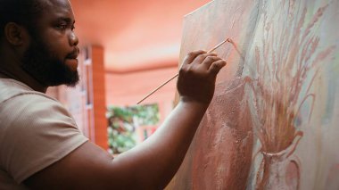 Erkek sanatçı suluboya ve palet ile modern bir sanat eseri çiziyor, sanat ve zanaat atölyesinde yeni eseri üzerinde çalışıyor. Genç ressam tuvalde kırsal bir başyapıt yaratıyor. Kamera B.