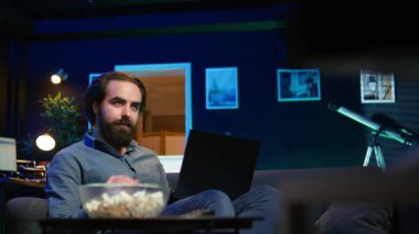 Dizüstü bilgisayarda çalışan adam dikkat dağıtmak için televizyon kanalını seçerken apartman dairesindeki kanepeye uzanmış. Uzaktan çalışan, dizüstü bilgisayardaki şirket görevlerini çözen, arka planda TV şovu, kamera B