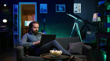 Teleworker evde televizyon izlerken dizüstü bilgisayarına e-posta yazıyor. Uzaktan kumandalı çalışan rahat bir apartman dairesinde çalışıyor, not defterine yazıyor, uzaktan kumandayla TV açıyor, B kamerasını kullanıyor.