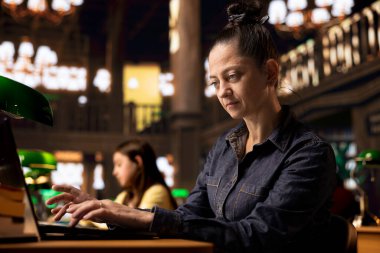 Sessiz bir kütüphane çalışma alanında dizüstü bilgisayarında yazı yazan mutlu kadın kütüphaneci, kendini kaynakları organize etmeye ve öğrencilere akademik yolculuklarında gurur ve uzmanlıkla rehberlik etmeye adadı..