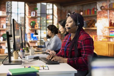 Ofisteki çağrı merkezi ajanı mikrofonla konuşuyor, müşteriyle iletişim kuruyor, adım adım rehberlik ediyor. Kadın, bilgisayar masasında müşterileri teknik çözümler üzerinden yürütüyor.